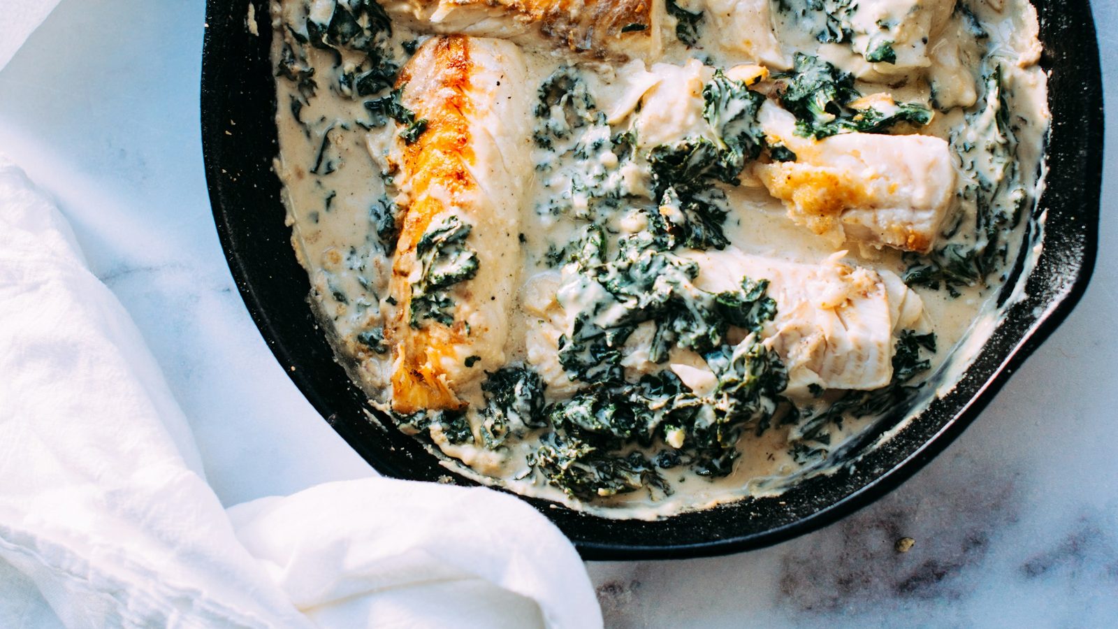 Fish fillets in a creamy kale sauce, in a cast iron skillet on a marble counter